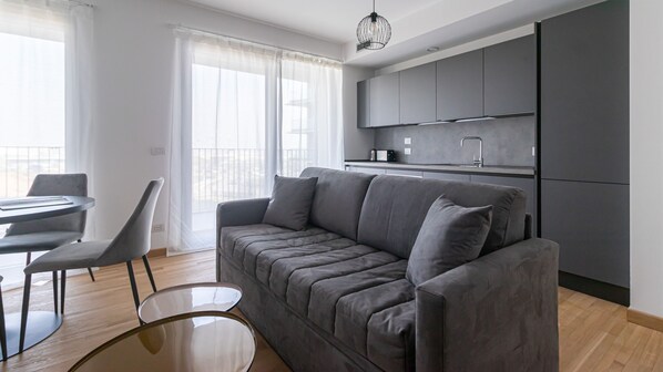 A modern living area with a grey sofa, a round glass coffee table, and a dining table with chairs. There is a kitchen with grey cabinets and a stainless steel sink.