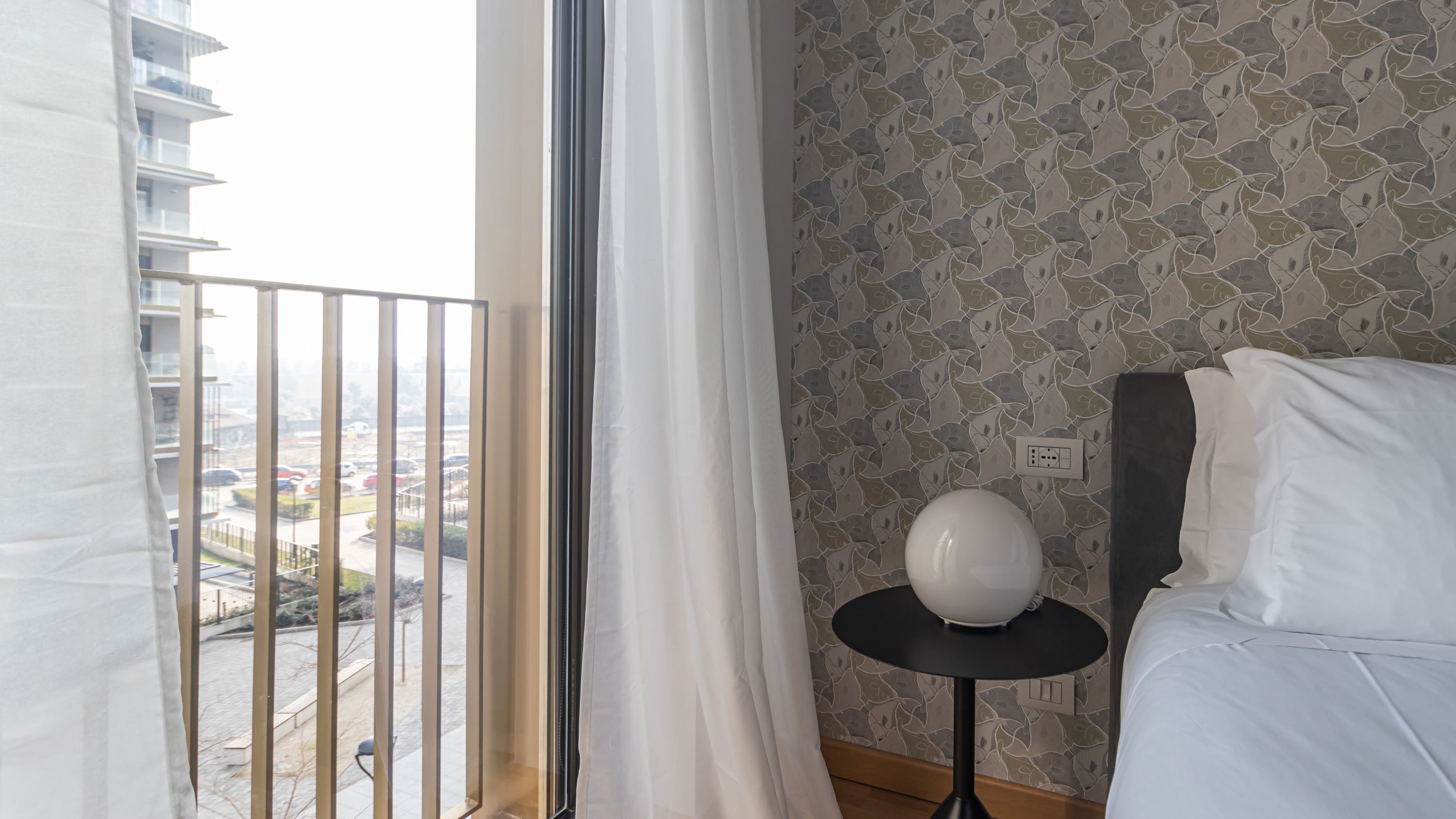 A hotel room with a bed, a round table with a white object on it, a window with a view of the city, and a patterned wall.