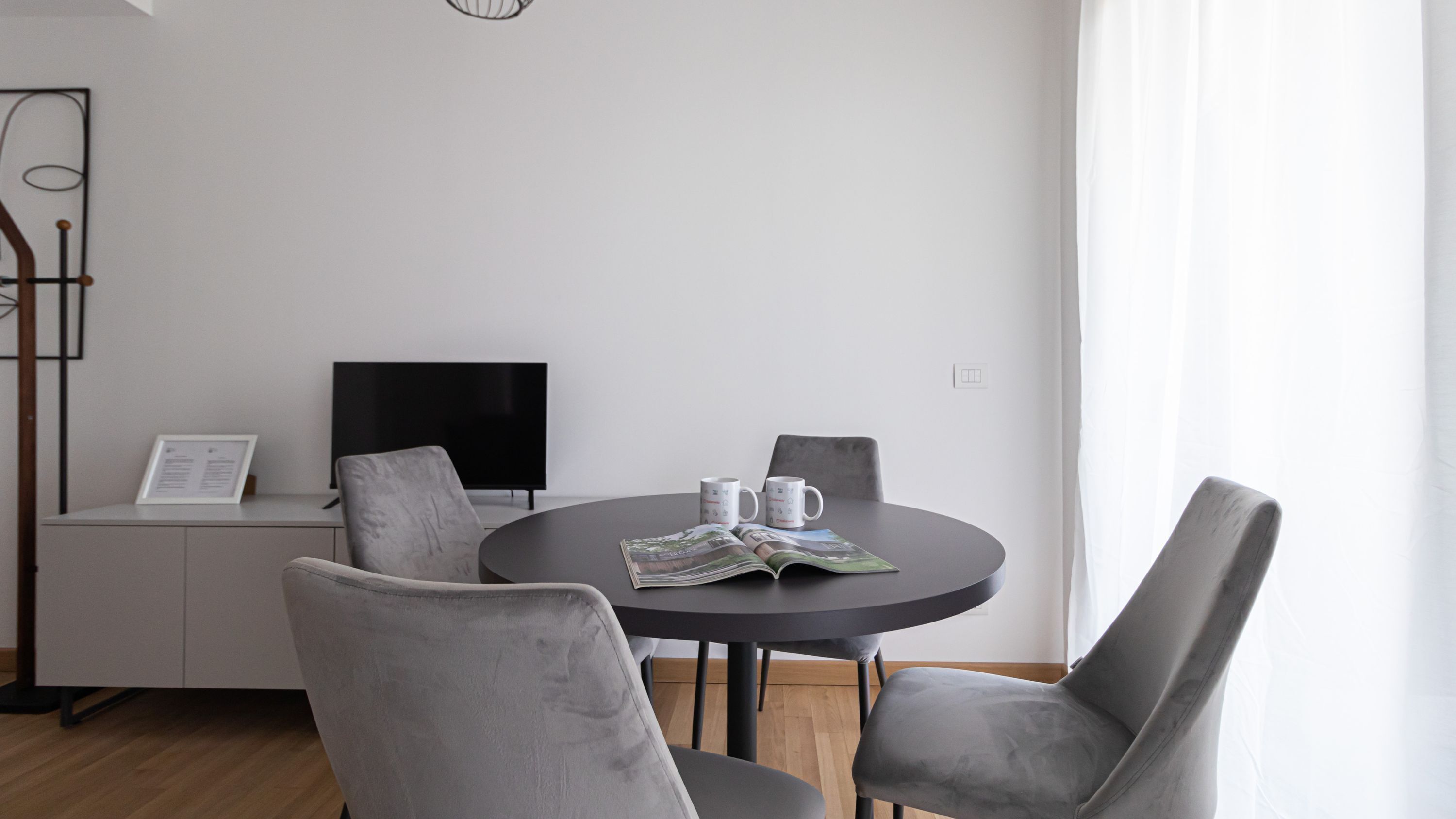 A modern living room with a round table, two grey armchairs, a flat-screen TV, and a magazine.