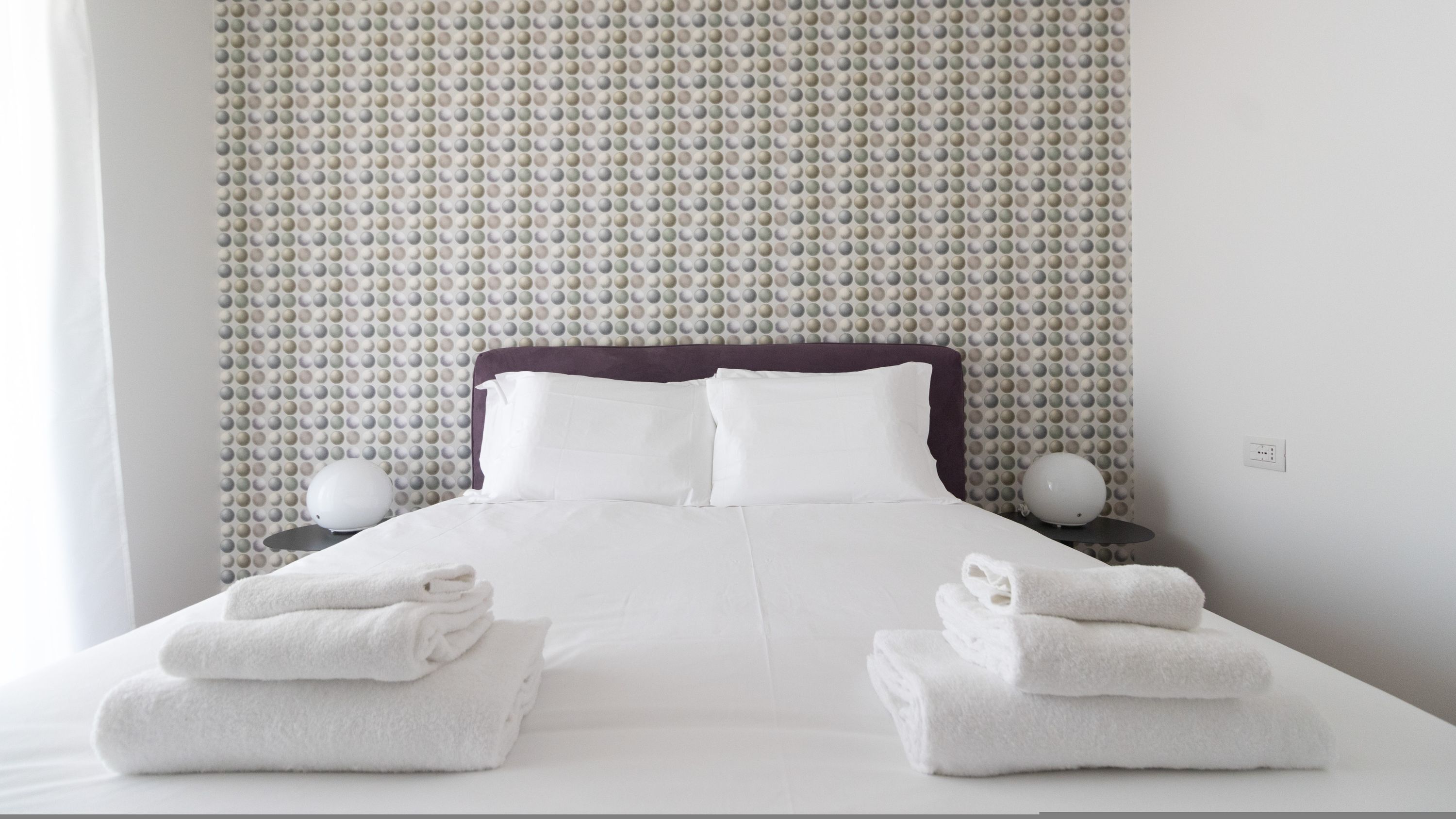 A neatly made bed with white linens and towels, a purple headboard, and a textured wall behind.