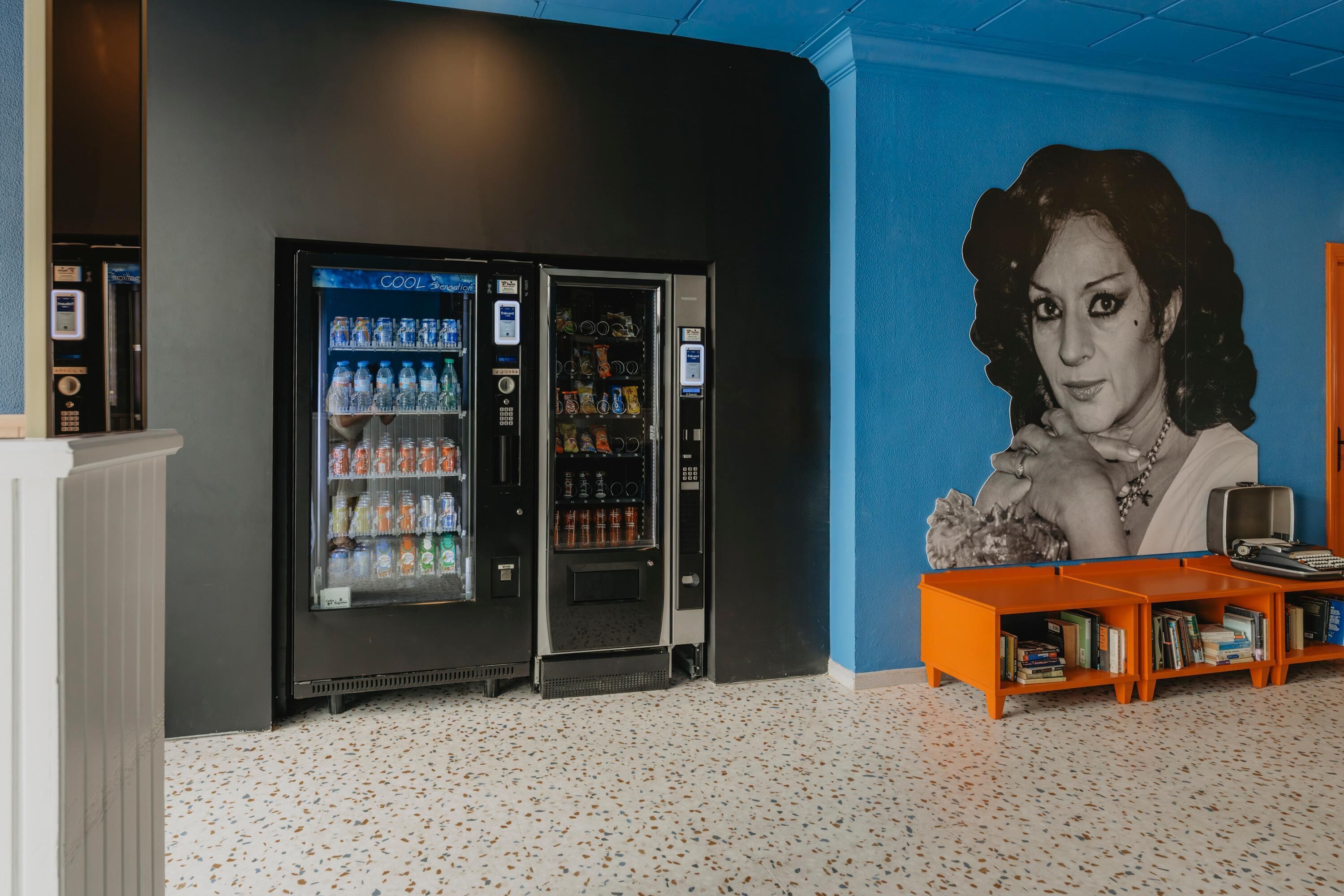 A room with two vending machines, a bookshelf, and a large portrait on the wall.