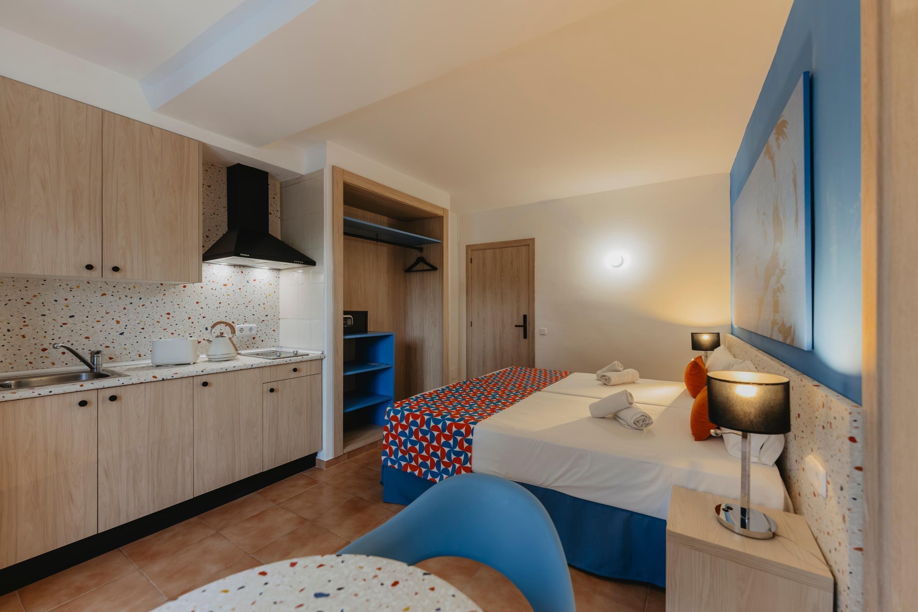 Una habitación de hotel compacta con kitchenette, una cama con colcha estampada, una mesita de noche con lámpara y una silla azul.