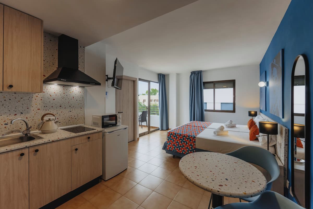 Una cocina compacta con lavamanos, hervidor y estufa, un pequeño comedor con mesa y sillas, y un dormitorio con cama y cortinas.