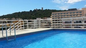 Una piscina con agua azul cristalina, una escalera metálica y una valla blanca. En el fondo, un edificio con varios balcones y una colina con árboles.
