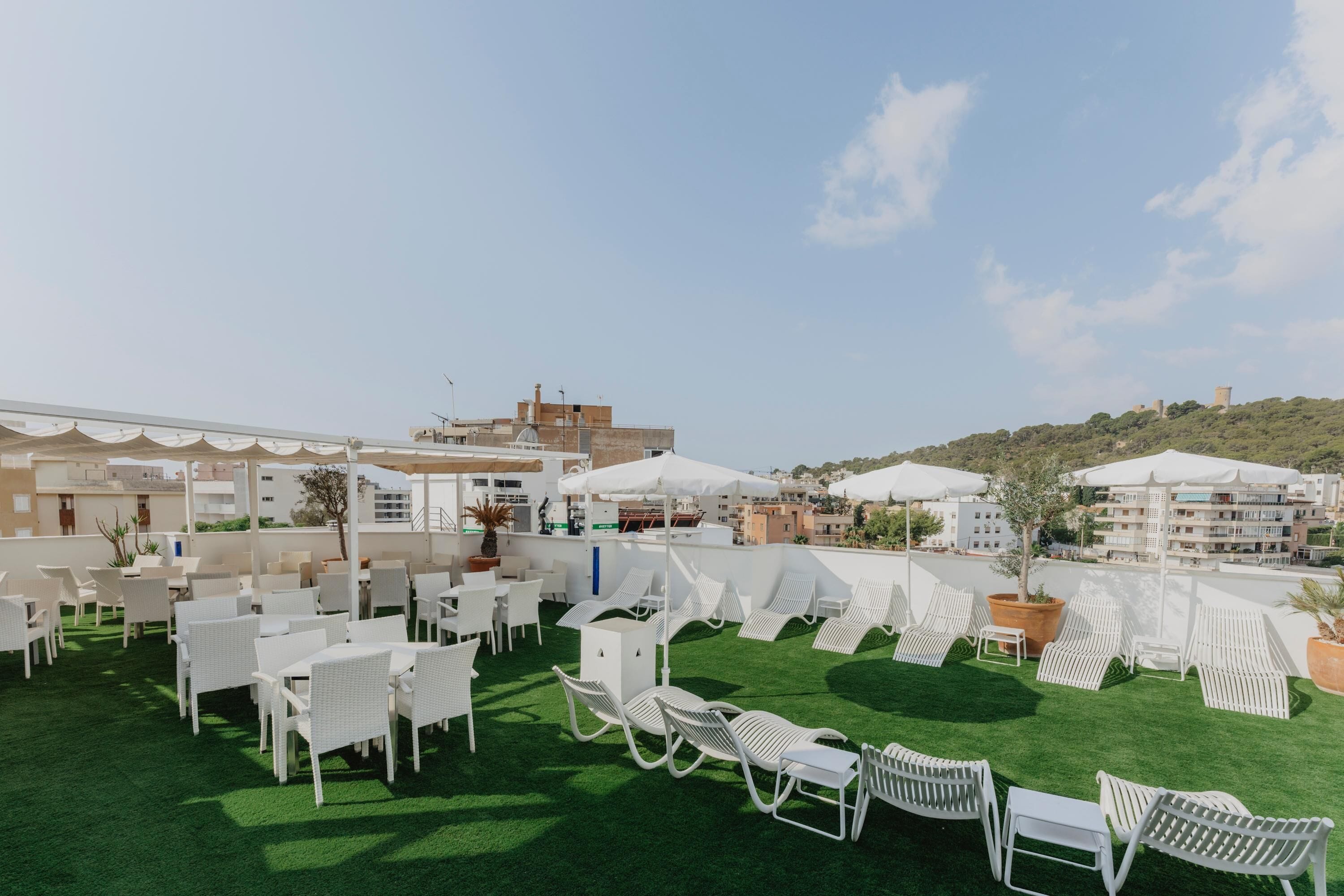 Terraza en la azotea con sillones blancos y mesas, rodeada de sombrillas, con vistas a un paisaje urbano.