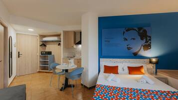 Una habitación de hotel con una cama, un escritorio con una silla, una kitchenette y un cuadro colgado en la pared.