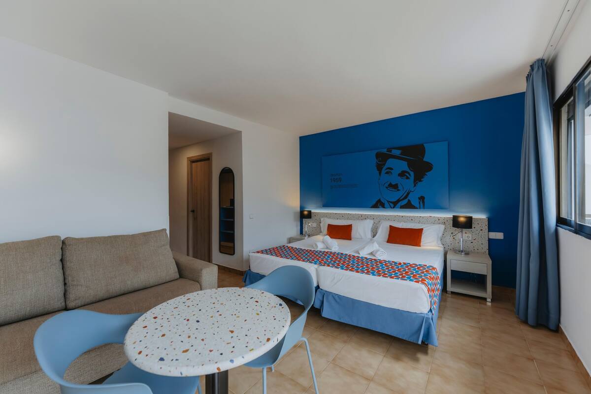 Habitación de hotel con cama, sofá, una mesita y una silla. Un cuadro cuelga en la pared sobre la cama.