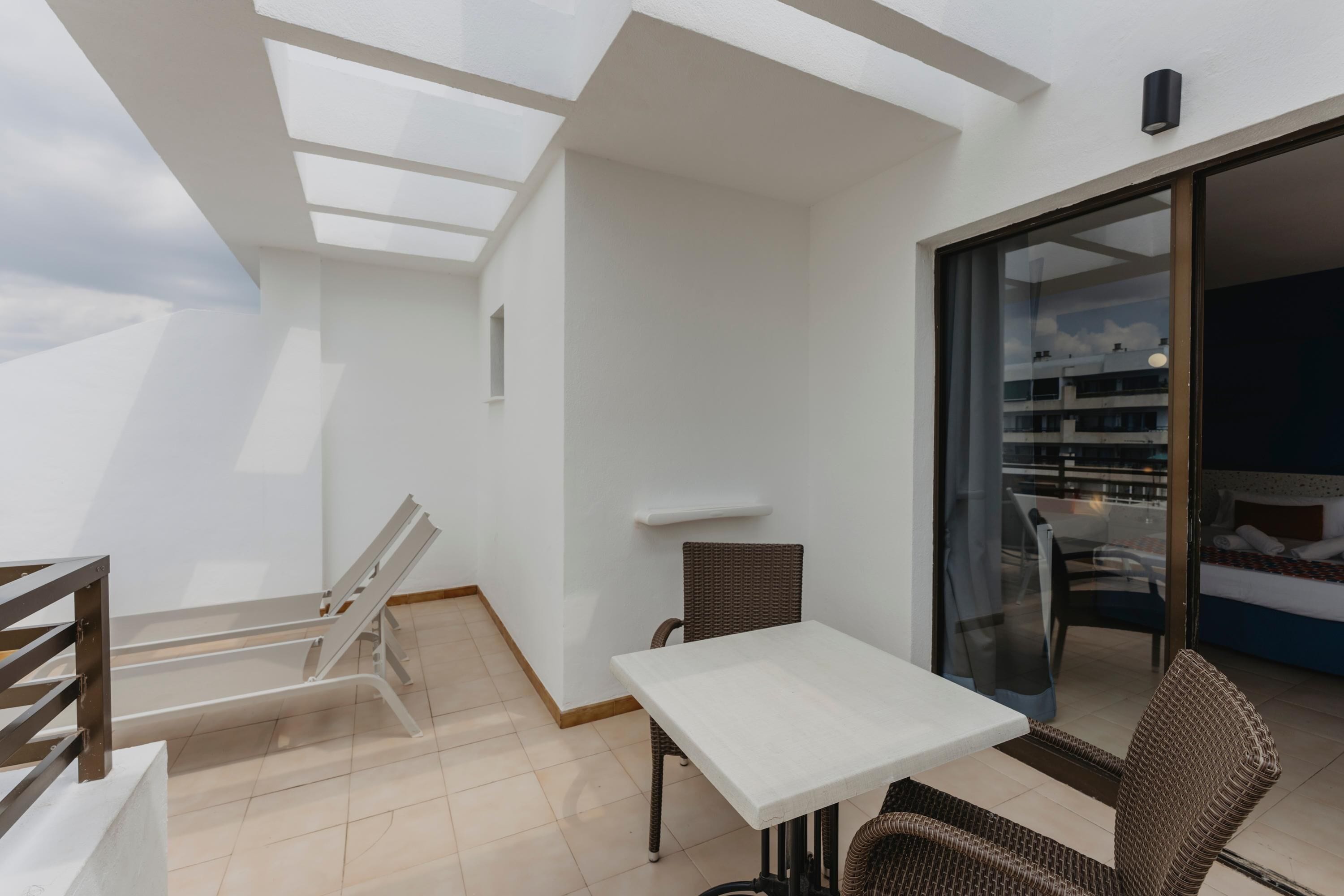 A balcony with a white table and wicker chairs, a glass door leading to a bedroom, and a skylight.