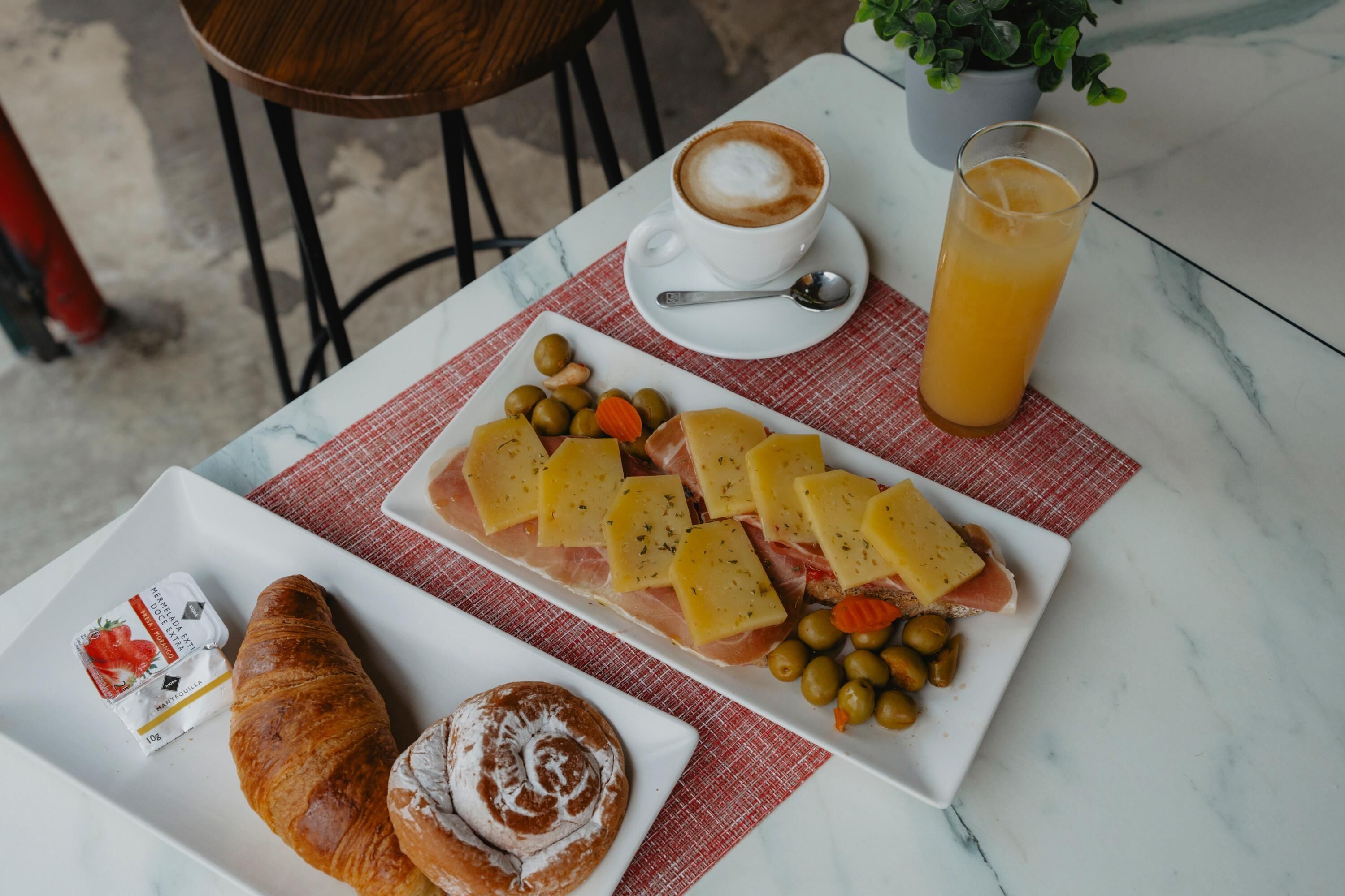 Una mesa con un croissant, queso, aceitunas y un vaso de zumo de naranja para el desayuno.