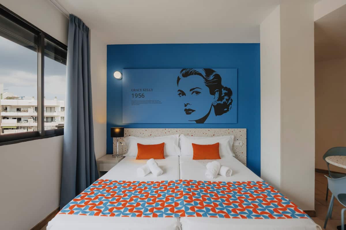 Habitación de hotel con una cama grande, dos almohadas, pared azul con un retrato y una ventana con cortinas.