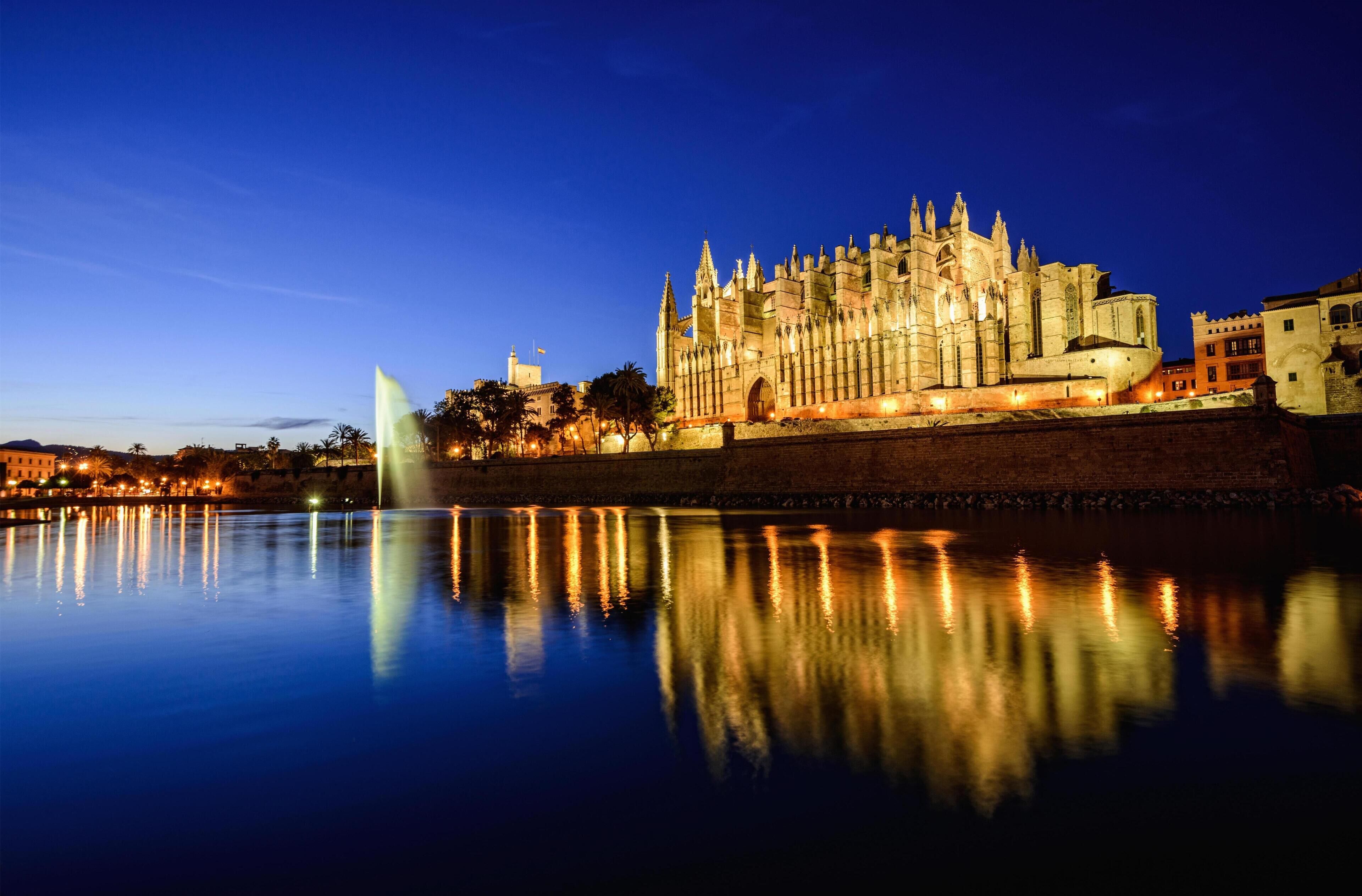 Una catedral histórica con arquitectura gótica, una masa de agua que se refleja en ella y una fuente a la izquierda.