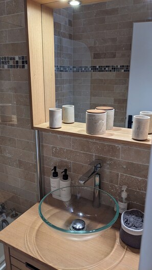 A bathroom with a glass sink, a wooden vanity, a mirror, and a shelf with toiletry items.