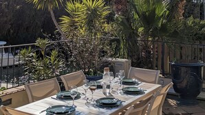 An outdoor dining area with a table set for two, surrounded by palm trees and other greenery. - On the water: XXL Terrace, Paddle, Bikes & A/C. New 2025 + 2 Secure Garages (Cogolin)