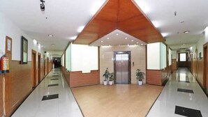 A long, well-lit hallway with a central elevator, flanked by wooden paneling and potted plants. - HOTEL O ONE HOTEL (Ghaziabad)