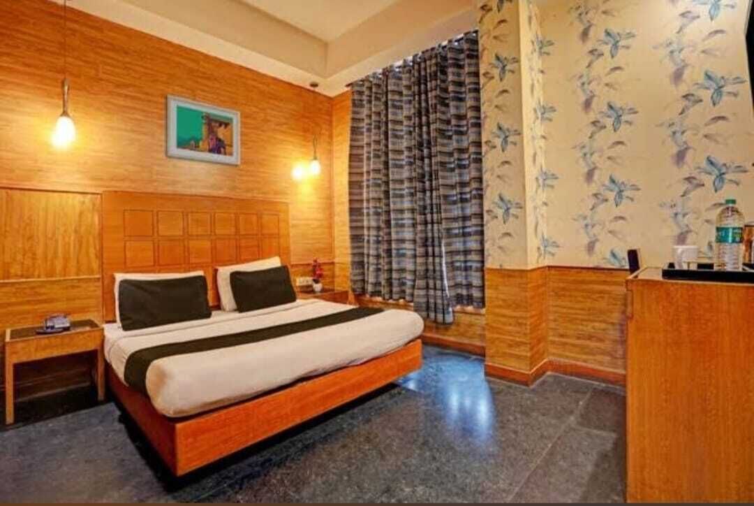 A hotel room with a wooden headboard, a bed with a white and black bedspread, a nightstand, a desk, and a patterned curtain.