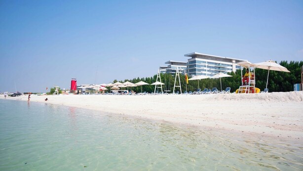 Ein Strand mit klarem Wasser, Liegestühlen, Sonnenschirmen und einem Gebäude im Hintergrund.