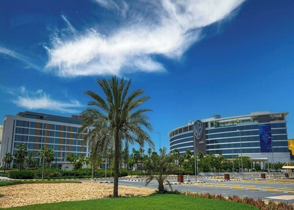 A modern hotel with a palm tree in the foreground, a clear blue sky, and a parking lot.