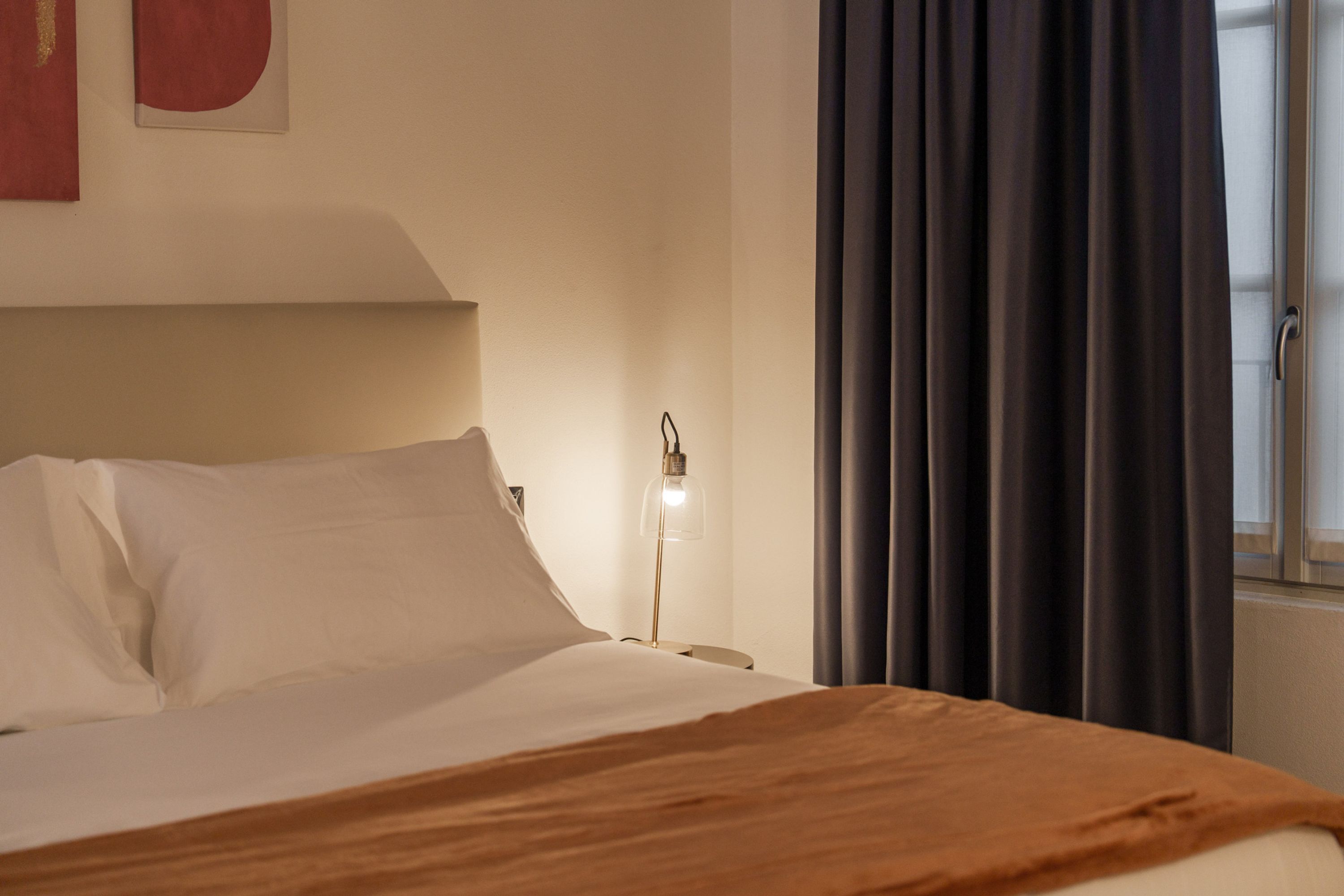 A neatly made bed with white linens and a brown throw blanket, a bedside lamp, and a window with dark curtains.