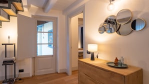 A well-lit hallway with a wooden staircase, a white door, a bedside table with a lamp, and a wall-mounted mirror. - Palazzo Odescalchi by Wonderful Italy - Apt 7 (Como)