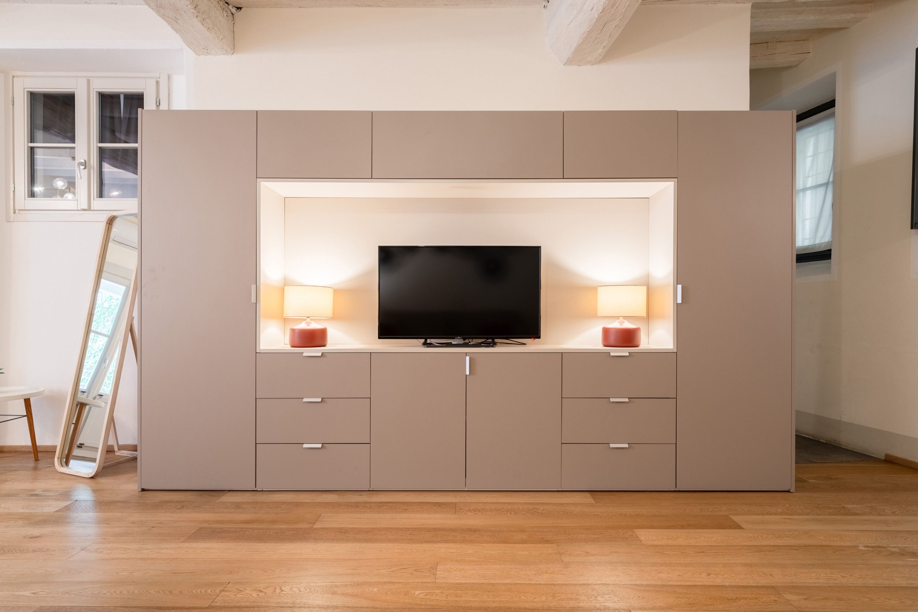 Un salon moderne avec un grand téléviseur posé sur un meuble intégré, un plancher en bois et une fenêtre avec des rideaux.