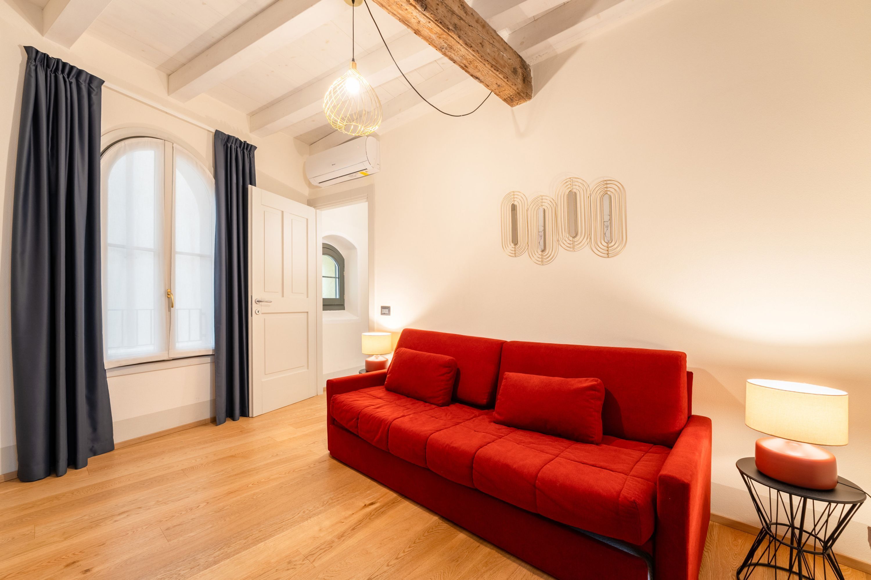 Ein modernes Wohnzimmer mit einem roten Sofa, Holzboden, einer Decke mit freiliegenden Balken, einem Fenster mit Vorhängen und einer Wandlampe.