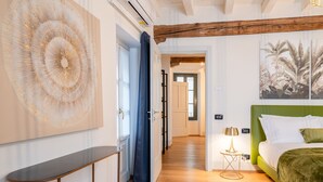 A hotel room with a wooden floor, a bed with green bedding, a wall-mounted artwork, and a small table with a lamp.