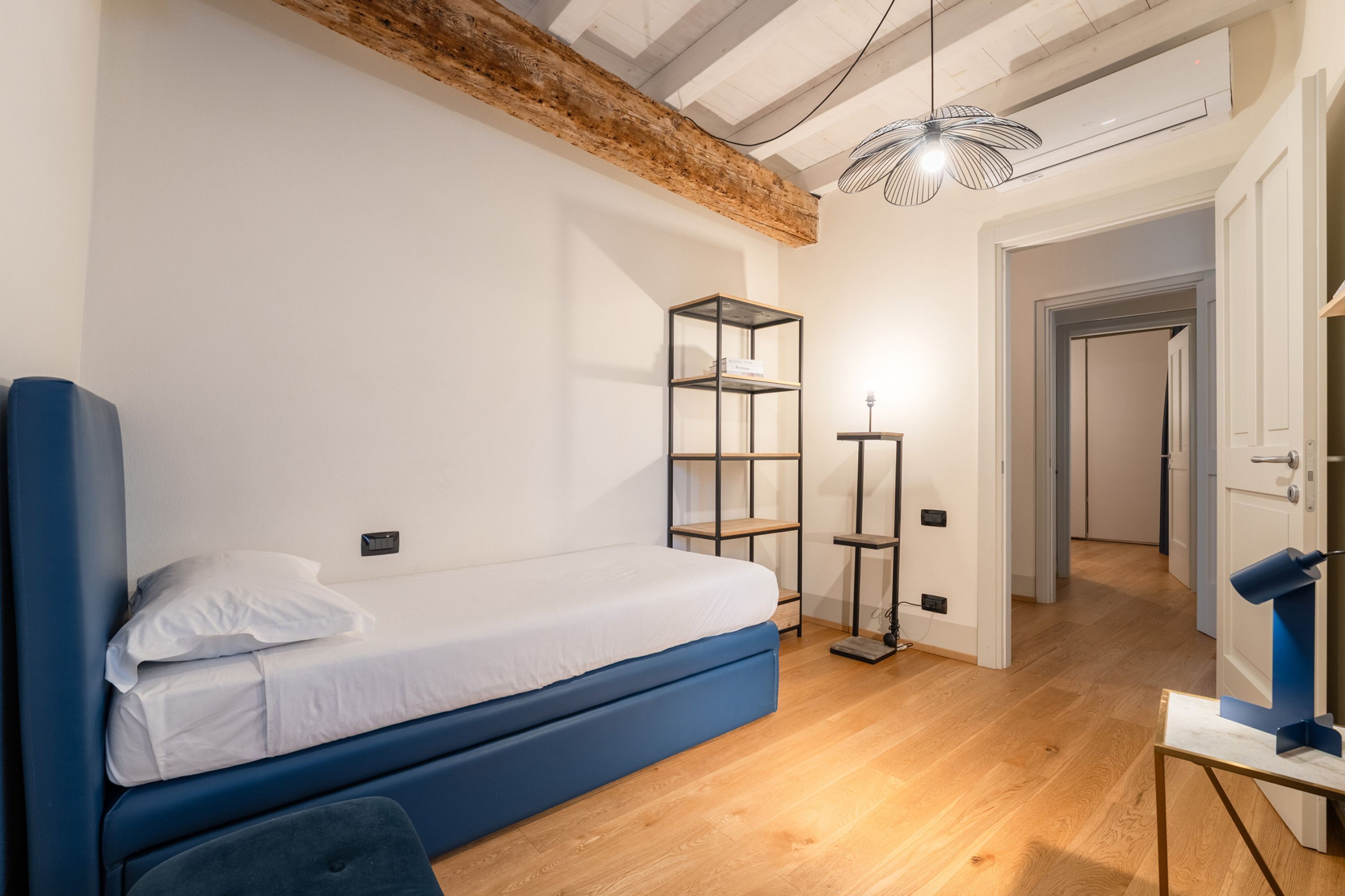 A bedroom with a blue bed, a wooden ceiling, a white wall, and a small table with a lamp.
