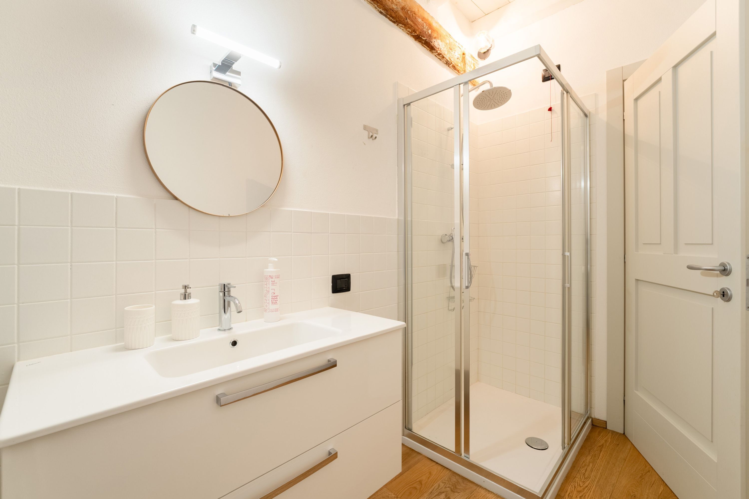 Une salle de bain moderne avec une douche à parois de verre, un lavabo blanc avec un miroir rond et un plancher en bois.