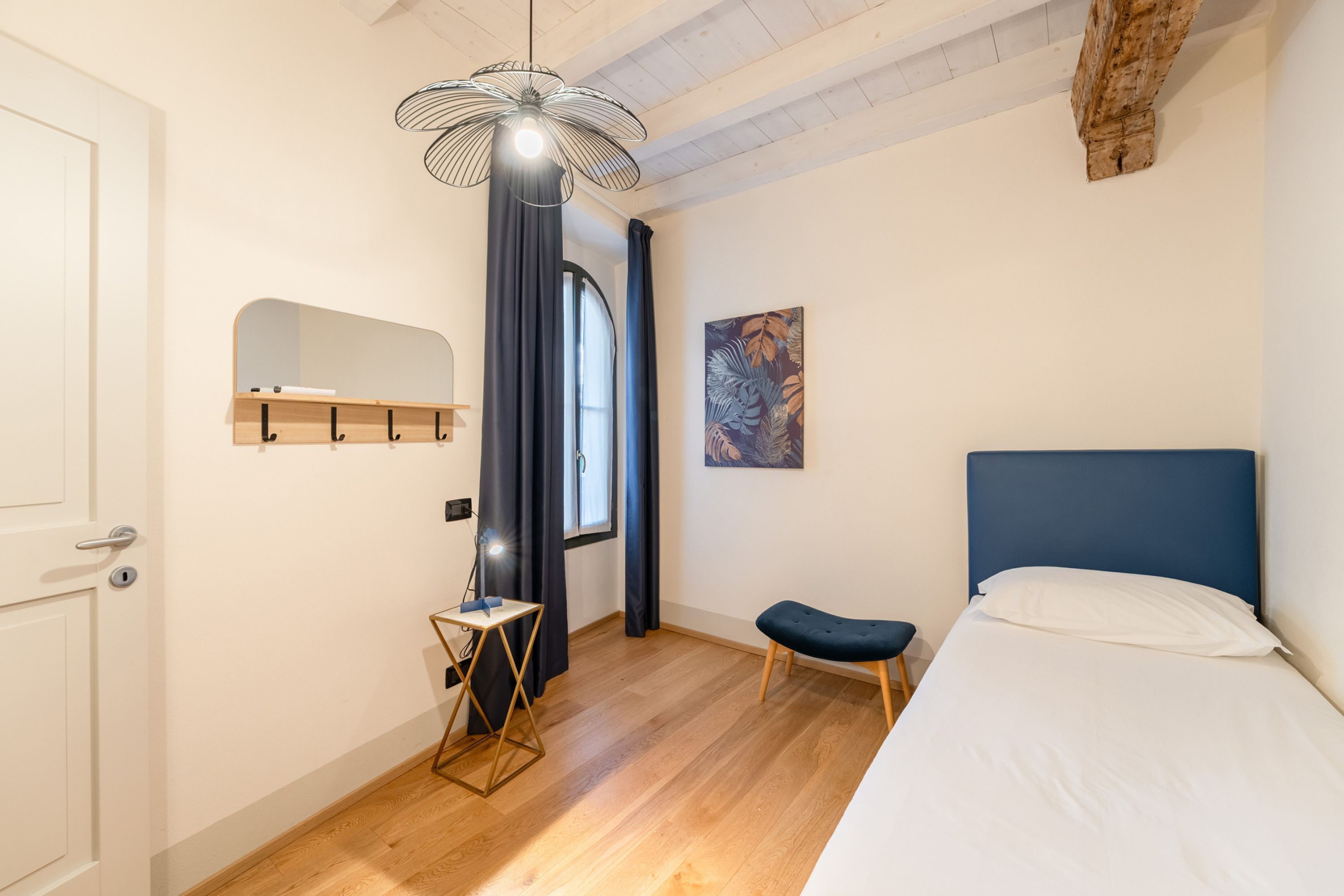 Une chambre à coucher comprenant un lit, un bureau, une chaise, un luminaire suspendu, une fenêtre avec des rideaux et un tableau accroché au mur.