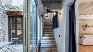 A narrow hallway with a wooden staircase leading to a second floor. A dining table and chairs are outside a glass door, and a sofa is visible in the adjacent room.