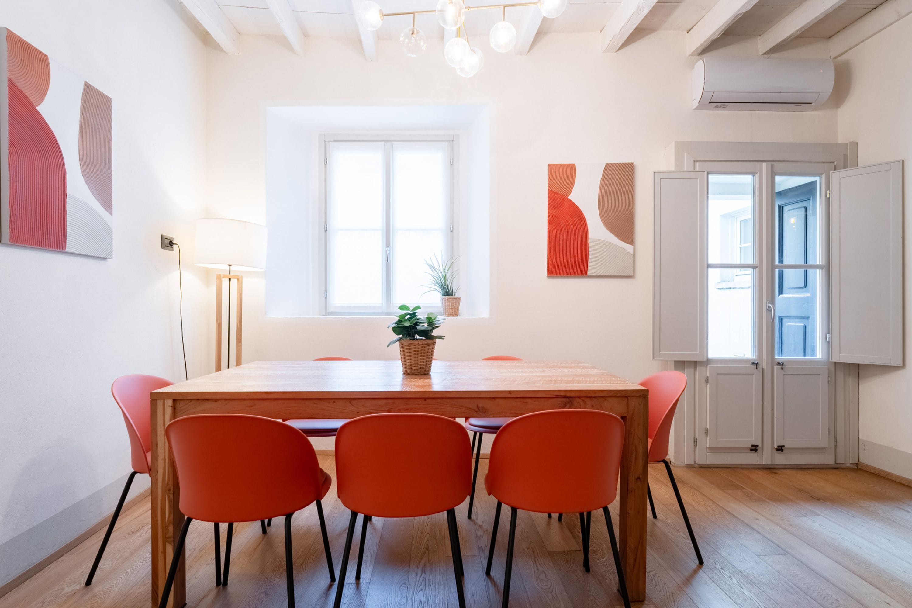 Een moderne eetruimte met een houten tafel, zes oranje stoelen, een potplant en abstracte wanddecoratie.