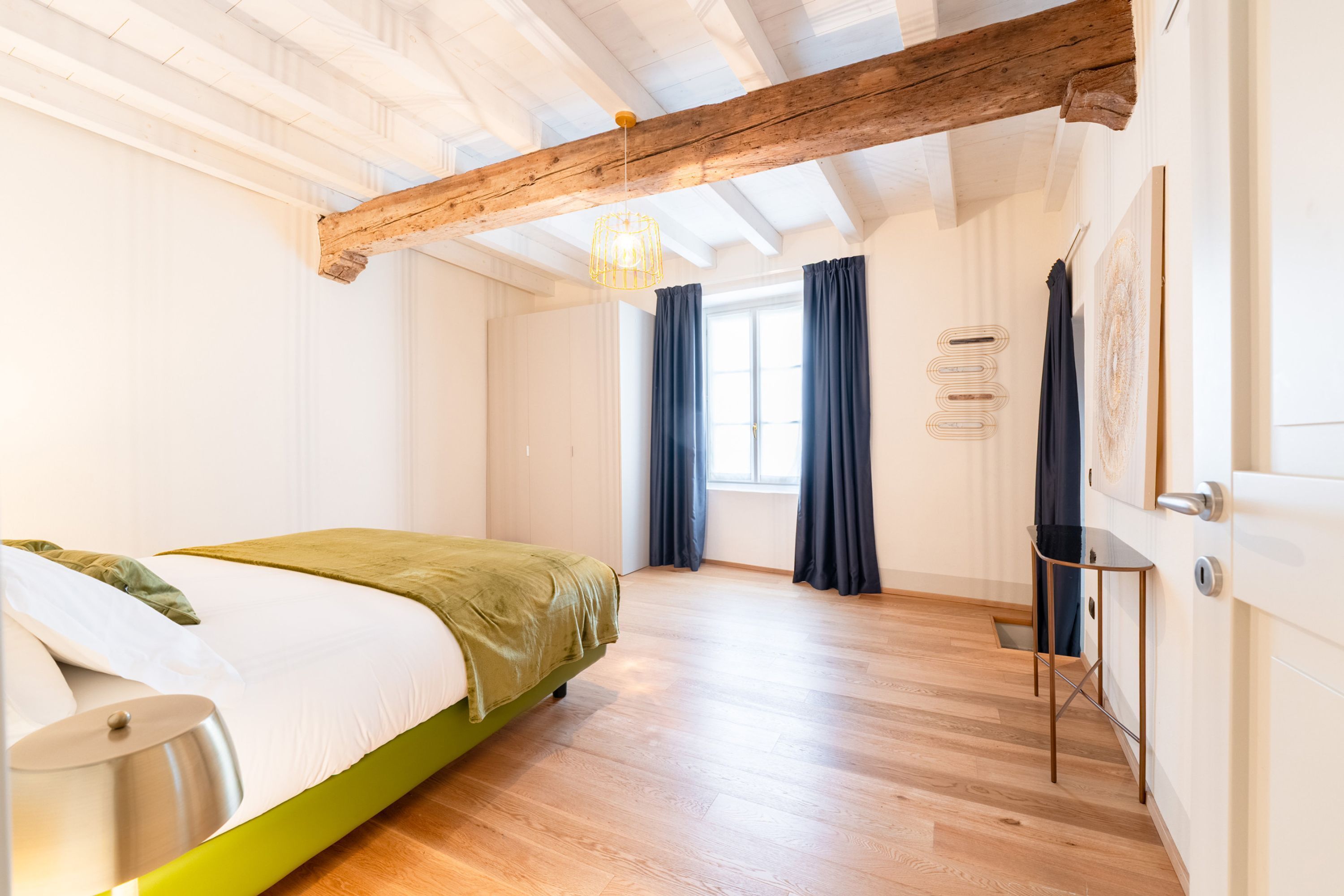 A modern bedroom with a wooden ceiling, a green bed, a bedside table, and a window with curtains.