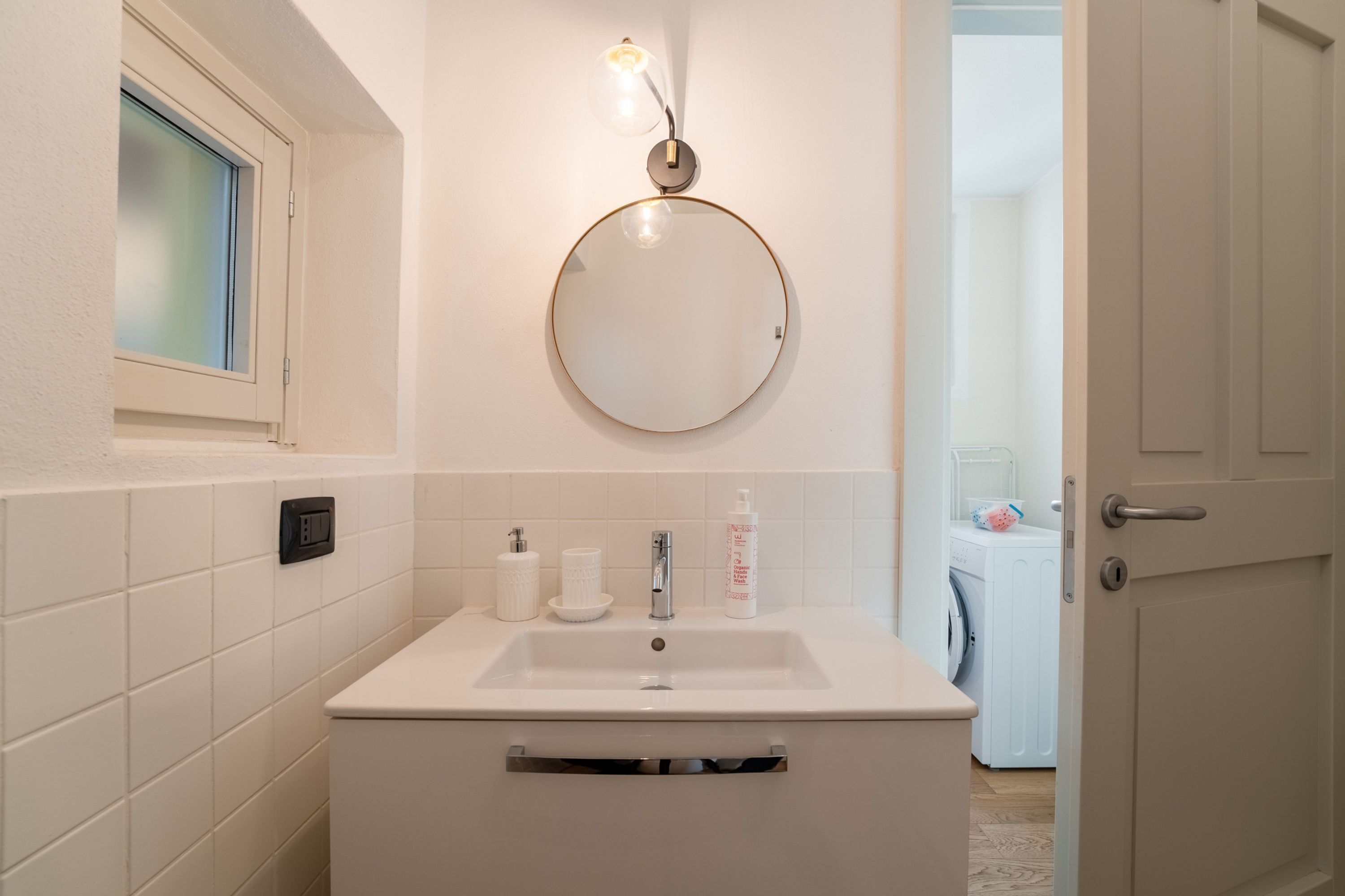 Une salle de bain avec un lavabo blanc, un miroir rond et une fenêtre à vitre dépolie.