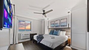 Un dormitorio con una cama, un ventilador de techo, un televisor montado en la pared, una ventana con vista al atardecer y obras de arte en la pared.