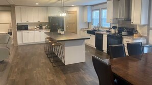 A modern kitchen with white cabinets, a central island with bar stools, a dining area with a wooden table, and a large window providing natural light. - Mountain Top Retreat with ATV Route Access (LaFollette)