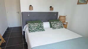 Une chambre à coucher avec un lit, des coussins à motifs verts, une tête de lit grise, une table de chevet avec un panier, et un panier fixé au mur.