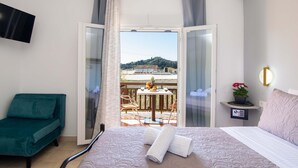 A hotel room with a bed, a green chair, a balcony with a table and chairs, and a view of a hill.