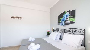 A hotel room with two beds, white linens, a decorative wall art of blue flowers, a small potted plant, and a wall-mounted light fixture.