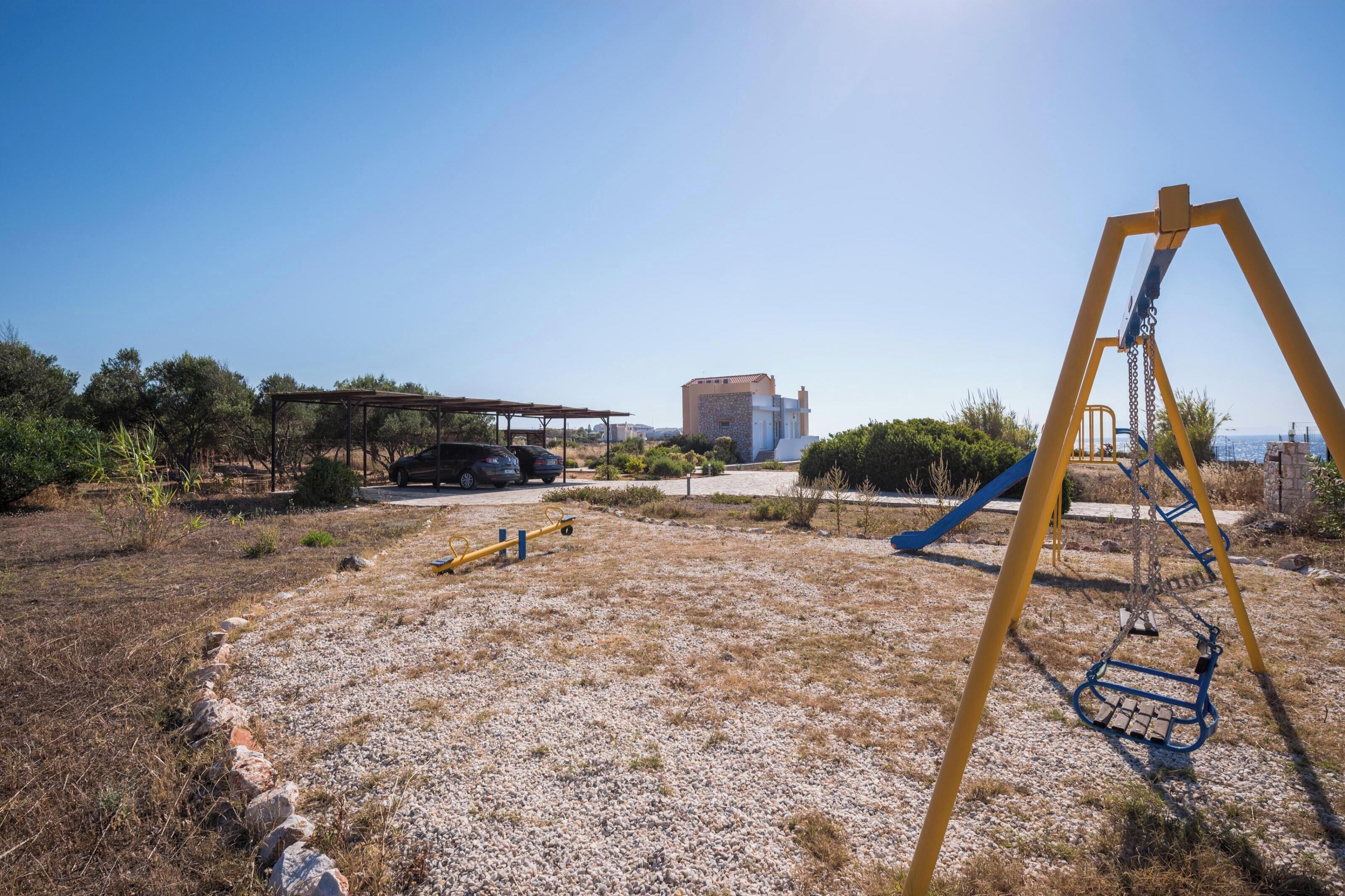 A playground with swings and a slide, surrounded by a gravel area and sparse vegetation.