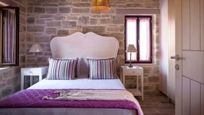 Un dormitorio con pared de piedra, cama blanca con almohadas a rayas, colcha morada y lámpara colgante.