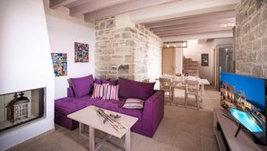 Un salón con un sofá morado, una mesa de centro y una pared de piedra. Hay una zona de comedor con una mesa y sillas, y un televisor que muestra una vista panorámica.