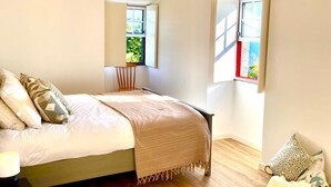 A bedroom with a wooden floor, a bed with a beige blanket, a bedside table, and a window with a view of greenery. - Liiiving in Azores - Casa de Foros (Lajes do Pico)