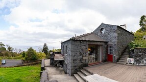 A stone building with a red door, a small outdoor seating area, and a staircase leading up to the entrance. - Liiiving in Azores - Casa de Foros (Lajes do Pico)