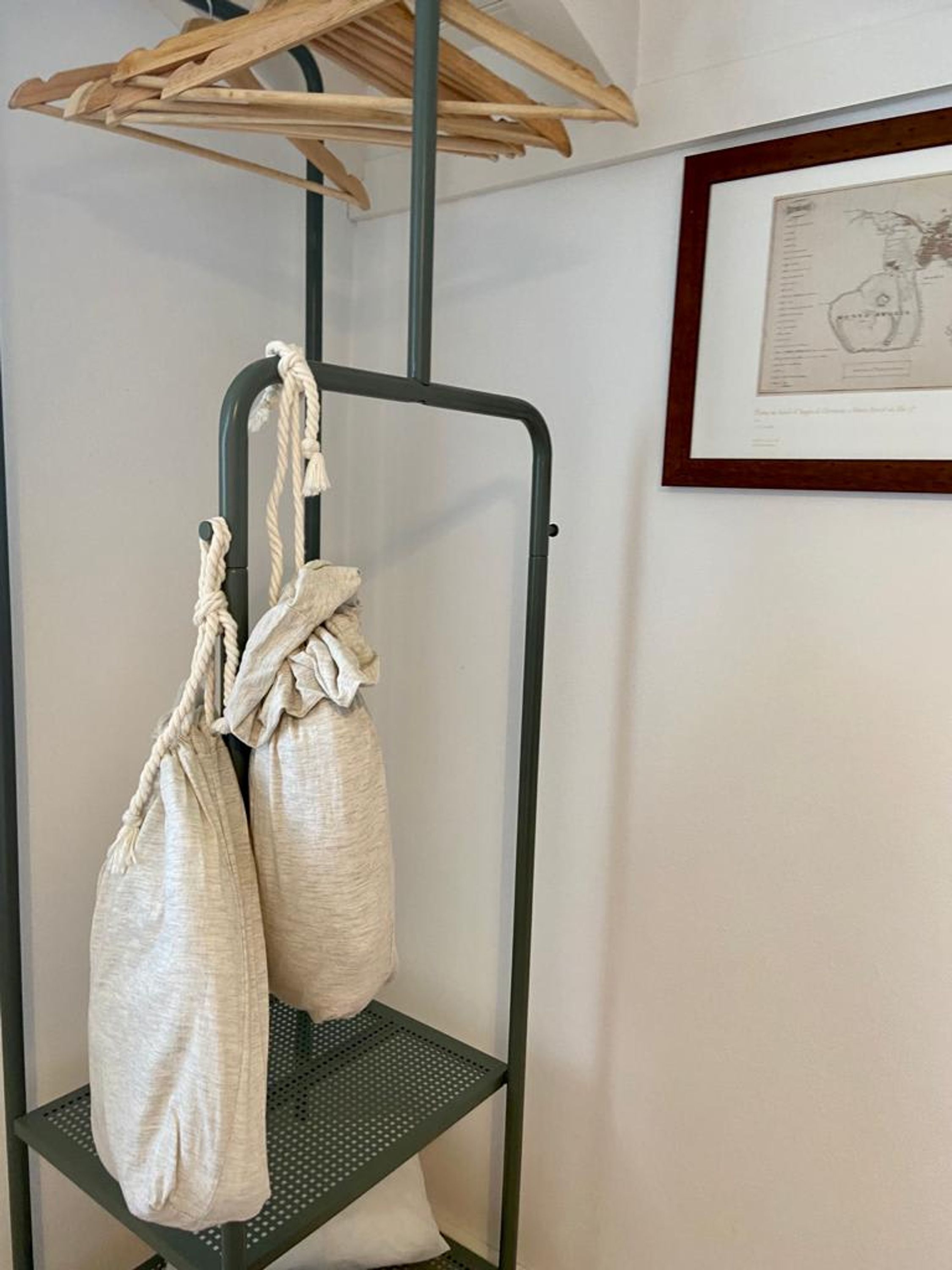 Un perchero de metal con dos bolsas colgadas, un cuadro en la pared y un tendedero de madera encima.