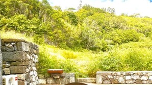 A wooden table with a green decorative plate in the center, set against a stone wall with a view of a lush green hillside. - Liiiving in Azores - Casa de Foros (Lajes do Pico)