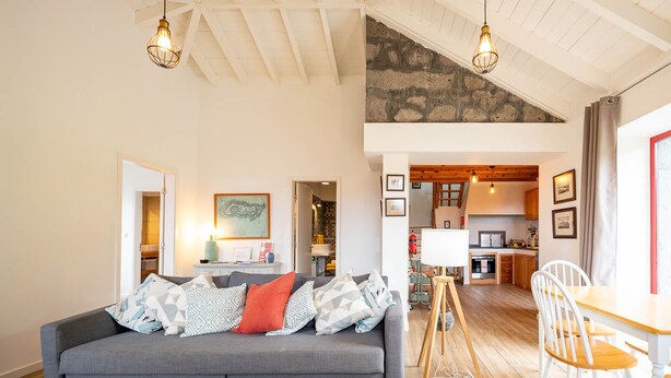 A modern living room with a grey sofa, wooden coffee table, and a dining area with a wooden table and white chairs. A kitchen is visible in the background. - Liiiving in Azores - Casa de Foros (Lajes do Pico)