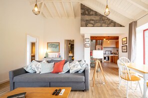 A modern living room with a grey sofa, wooden coffee table, and a dining area with a wooden table and white chairs. A kitchen is visible in the background. - Liiiving in Azores - Casa de Foros (Lajes do Pico)