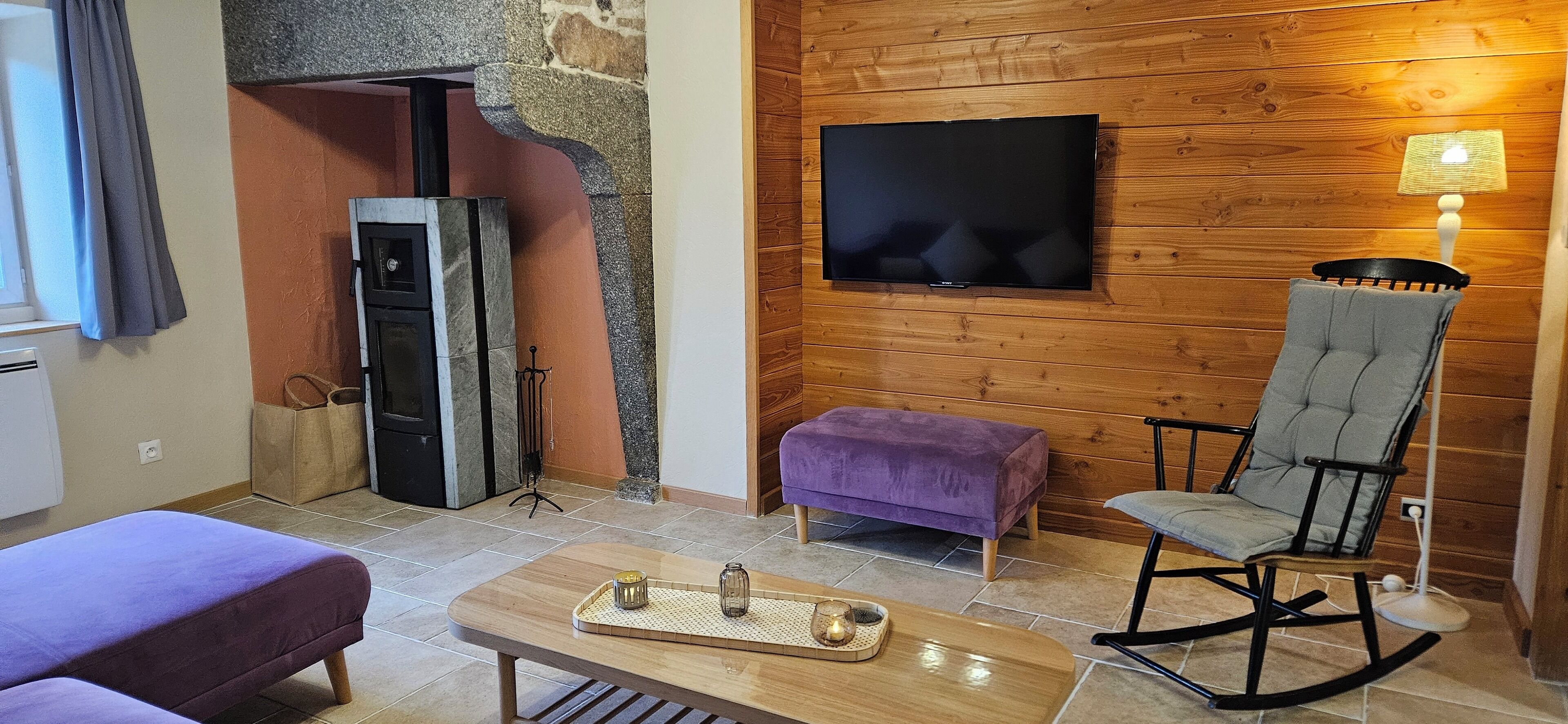 Ein gemütliches Wohnzimmer mit Kamin aus Stein, einem an einer Holzwand montierten Flachbildfernseher, einem Schaukelstuhl und einem violetten Hocker.