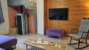Una sala de estar acogedora con chimenea de piedra, un televisor de pantalla plana montado en una pared de madera, un sillón mecedor y un reposapiés púrpura.