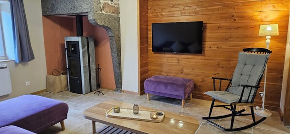 A cozy living room with a stone fireplace, a flat-screen TV mounted on a wooden wall, a rocking chair, and a purple ottoman. - House on the river (La Bresse)